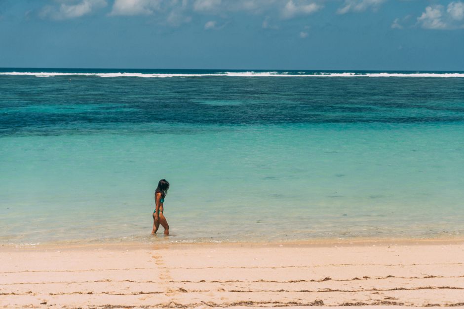 A woman walks along a tranquil beach with turquoise waters, enjoying a peaceful summer day.