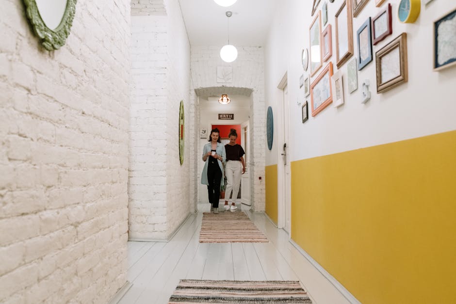 Two women walking through a stylish corridor adorned with art frames on white brick walls.