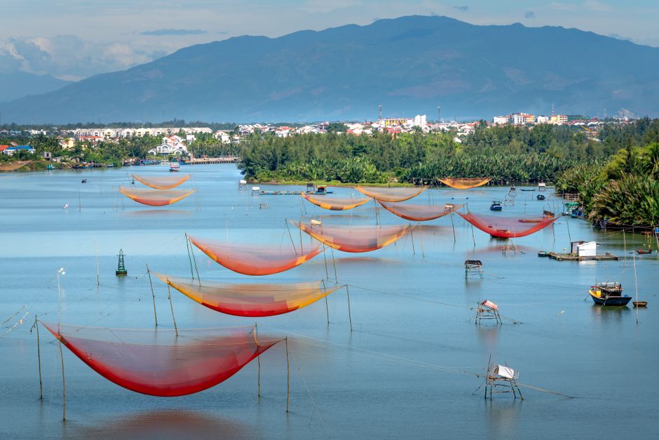 Vibrant fishing nets stretch across a serene lake near a lush town with distant mountains.