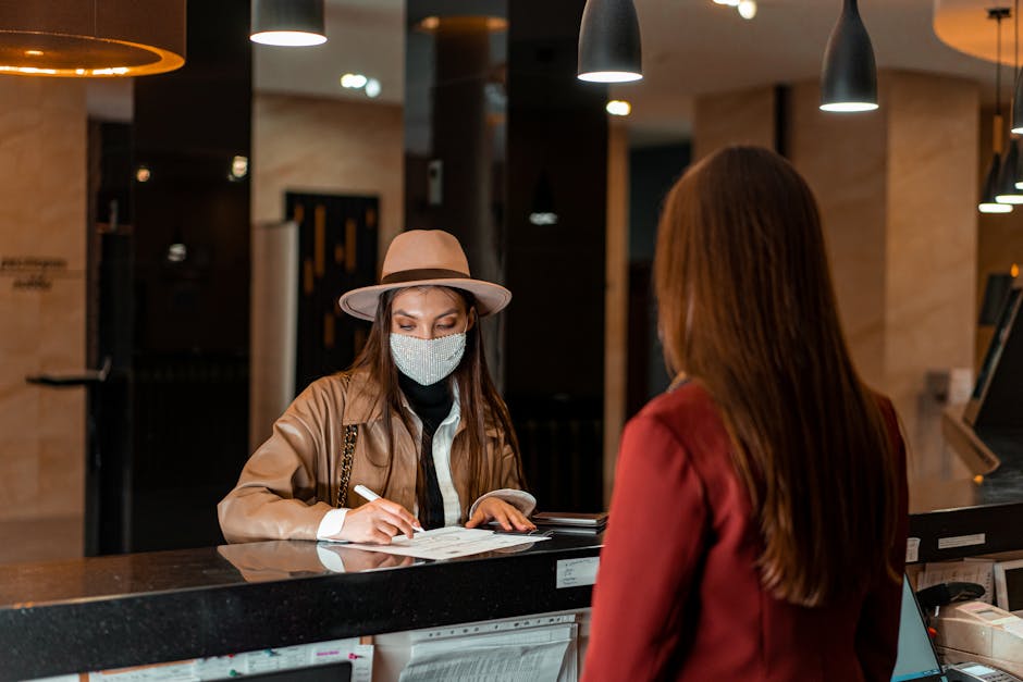 A woman in a hat and face mask checks in at a hotel reception, interacting with a receptionist.