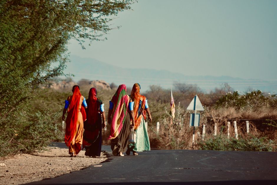 Five women in vibrant traditional attire walk down a rural road in India.
