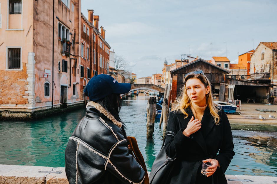 Two women conversing by a Venetian canal with historic architecture in view.