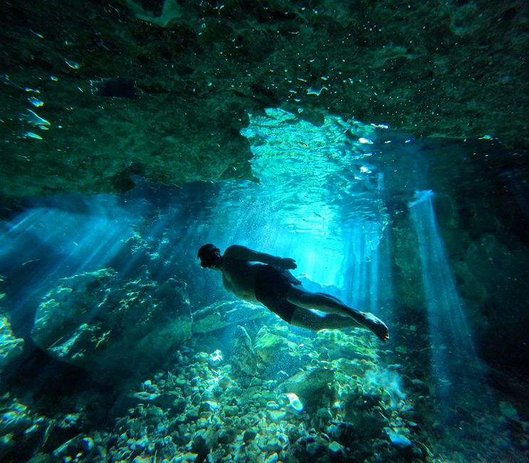 Freediver swimming through a sunlit underwater cave with dazzling blue waters.