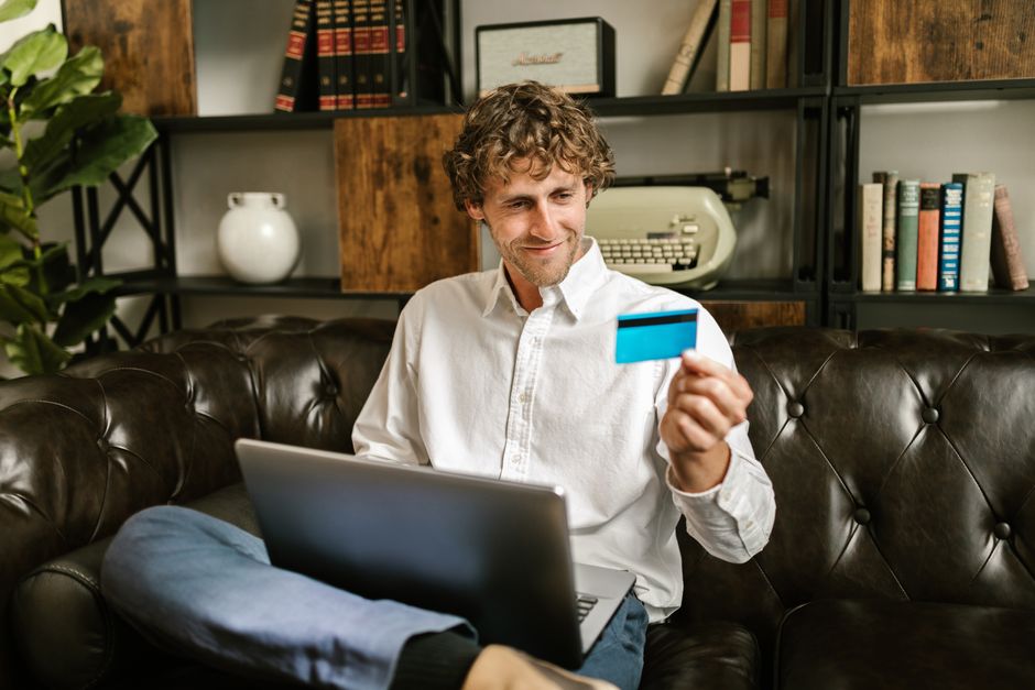 A man sits on a sofa, smiling while holding a credit card with a laptop on his lap, indoors.