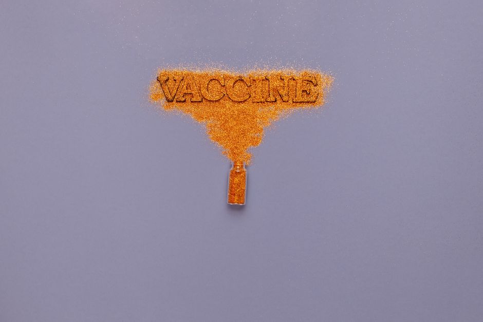 Orange glitter powder spreading into the word 'Vaccine' from a vial on a gray background.