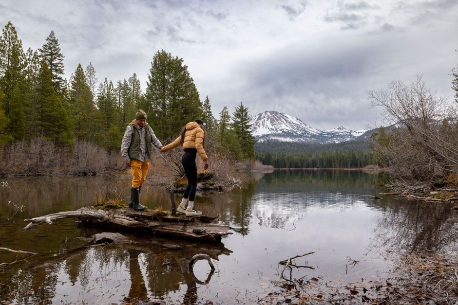 A couple explores nature, standing on driftwood over a serene lake with mountains in the background.