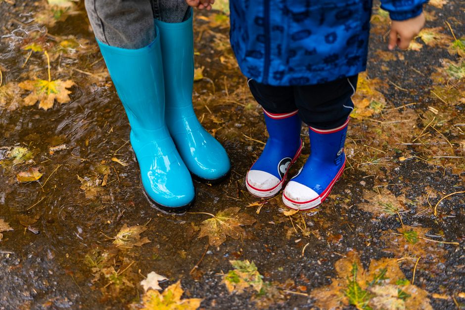 Two children wearing bright rubber boots splash in autumn puddles covered with fallen leaves.