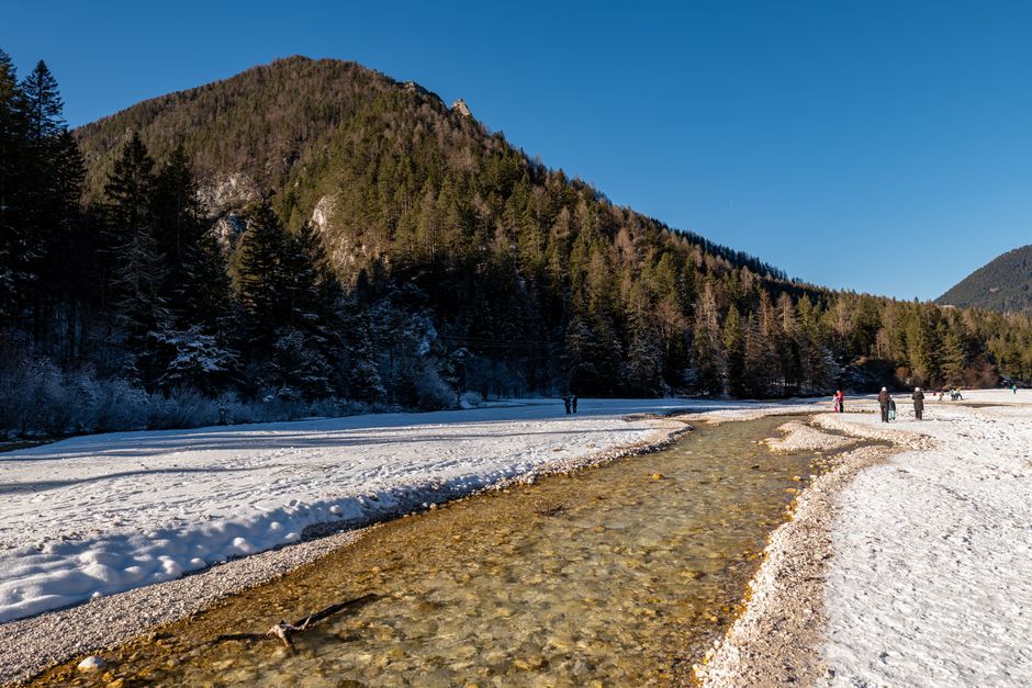A tranquil winter scene featuring a river in Kranjska Gora, Slovenia, surrounded by snowy landscapes and forested mountains.